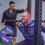 Muscular sportsman in activewear squatting with heavy barbell with effort while listening trainer in gym